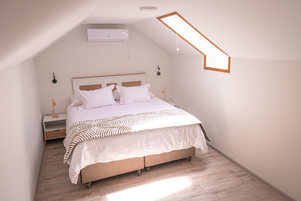 A well-lit bedroom features a spacious bed with neatly arranged white linens and decorative pillows. An air conditioning unit is mounted on the wall, and warm light fixtures flank the bed. A skylight brings natural light, enhancing the minimalist decor.
