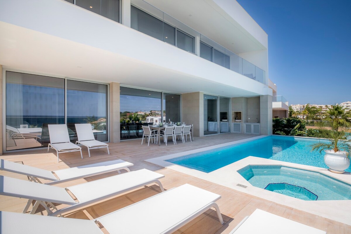 A modern outdoor area features a private swimming pool accompanied by a Whirlpool. Several white loungers are positioned on a wooden deck, with a dining table visible nearby under the overhang. Expansive glass doors provide a clear view of the sea and outdoor space.
