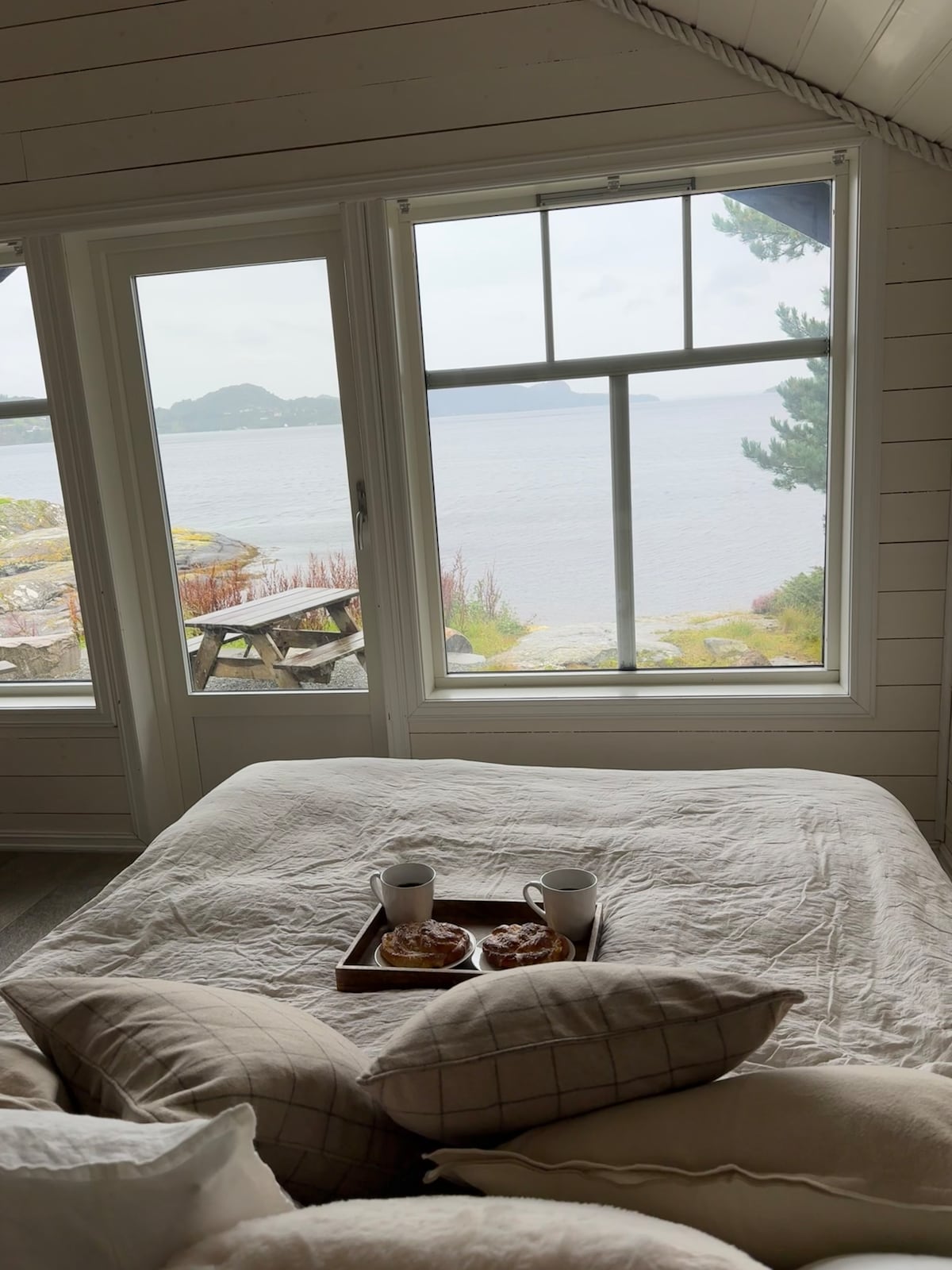 A spacious bed is positioned near large windows that frame views of the Fanafjorden. On the bed, a tray holds two mugs and pastries. Natural light fills the room, complemented by a serene atmosphere created by the surrounding landscape.