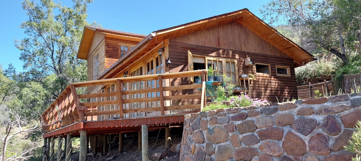 A wooden cabin with a welcoming deck is positioned on elevated stilts, surrounded by natural landscaping. Large glass windows allow ample sunlight into the interior space. The sturdy stone wall provides a rustic contrast to the wooden structure, contributing to the serene outdoor environment.