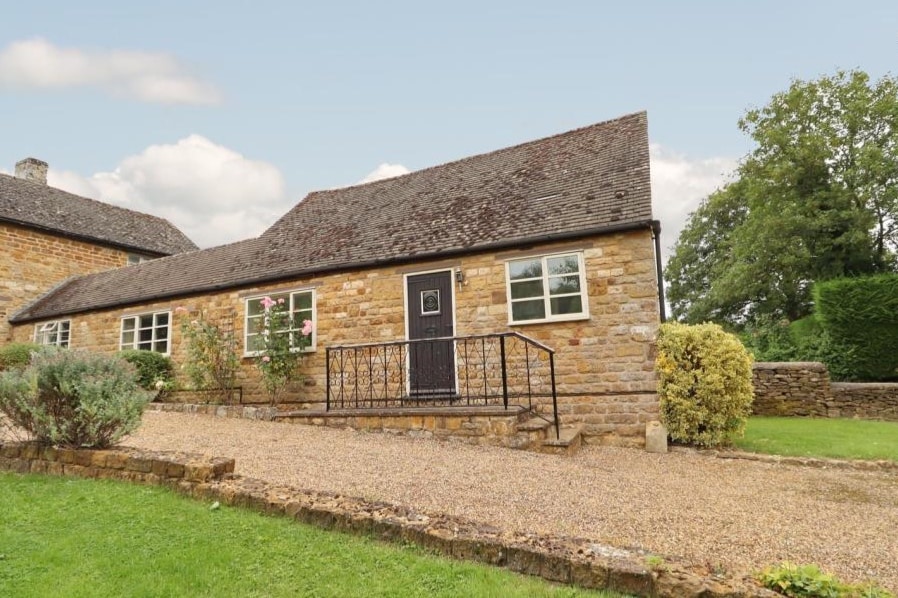 A traditional stone cottage is featured with a gravel pathway leading to the entrance. The cottage has large windows framed in dark wood and a simple, black door. Lush greenery surrounds the home, including shrubs and a neatly trimmed lawn.