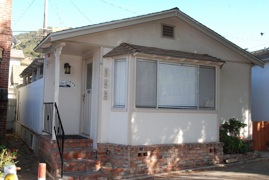 A single-story cottage features a light-colored exterior and brick detailing around the base. Large windows allow natural light into the space, while a small porch with a railing provides access to the entrance. A pathway leads to the door, surrounded by modest landscaping.