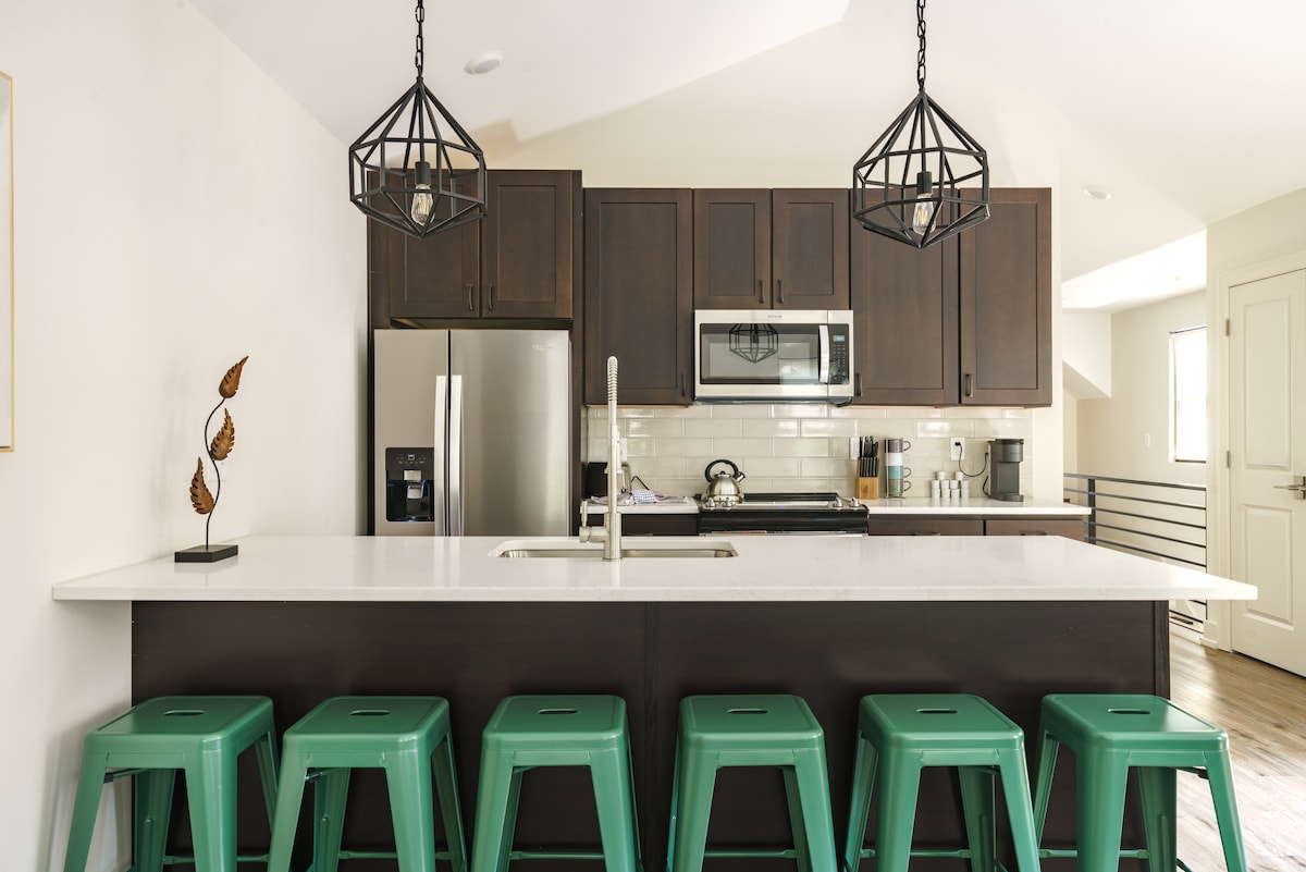 A modern kitchen is featured with dark wood cabinetry and stainless steel appliances. A spacious island with a white countertop is surrounded by six teal stools. Decorative light fixtures hang above, complementing the overall design while natural light enters from a nearby window.