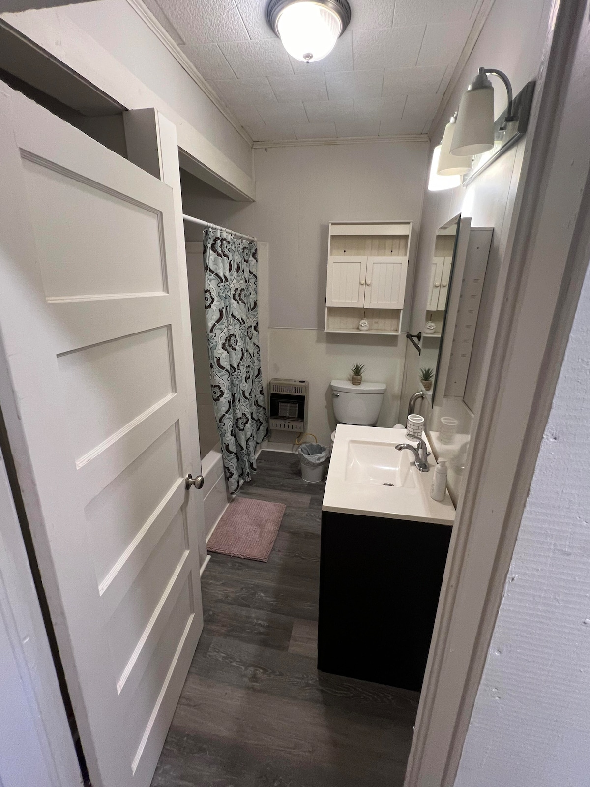 The bathroom is designed with light-colored walls and a dark vanity. A shower curtain with a floral pattern adds a touch of decor. A shelving unit is mounted above the toilet, providing storage. A soft rug lies in front of the sink area, complementing the overall layout.