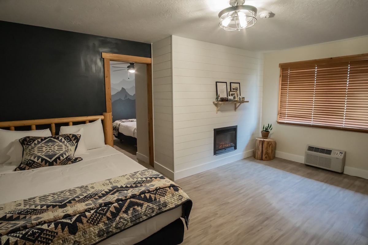 A well-appointed bedroom features a spacious bed with a patterned blanket, complemented by a cozy fireplace on one wall. Natural light enters through a window with wooden blinds, illuminating the room's neutral tones and enhancing its comfort.