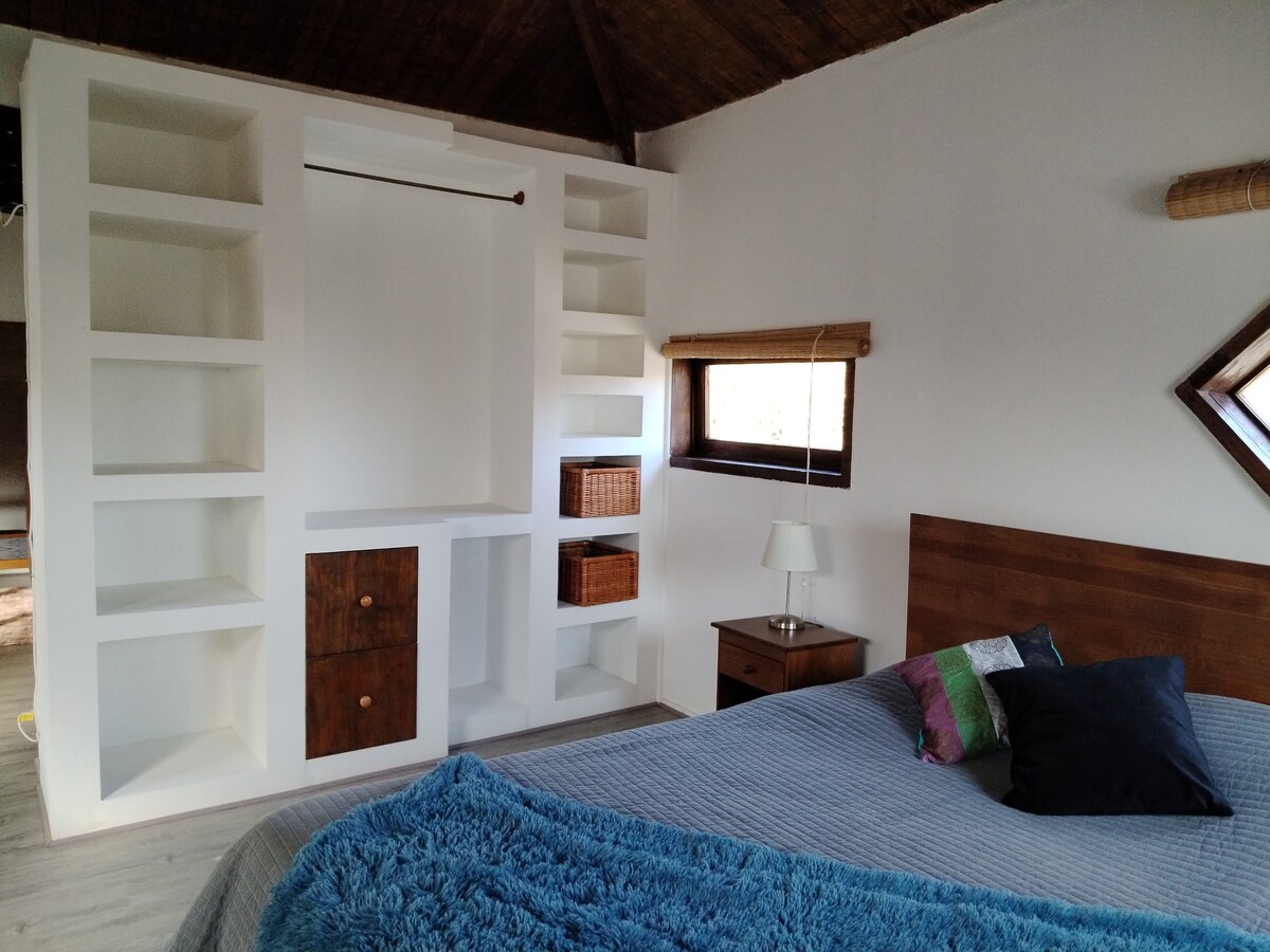 A spacious bedroom showcases a double bed with a textured blanket and decorative pillows. Built-in shelving provides ample storage, featuring woven baskets. A bedside table with a lamp sits next to the bed. Natural light filters through a window, adding warmth to the room.