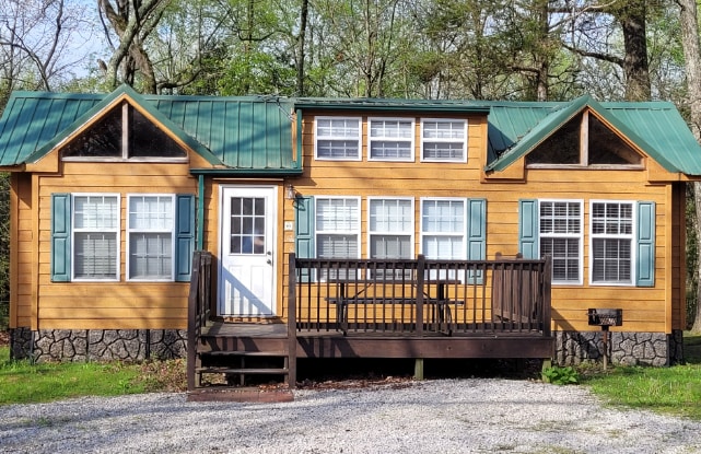 A charming cabin featuring a wooden exterior with a sloped green metal roof. Large windows provide ample natural light, and the front porch is accessible via a wooden railing. Surrounding greenery complements the inviting entrance.