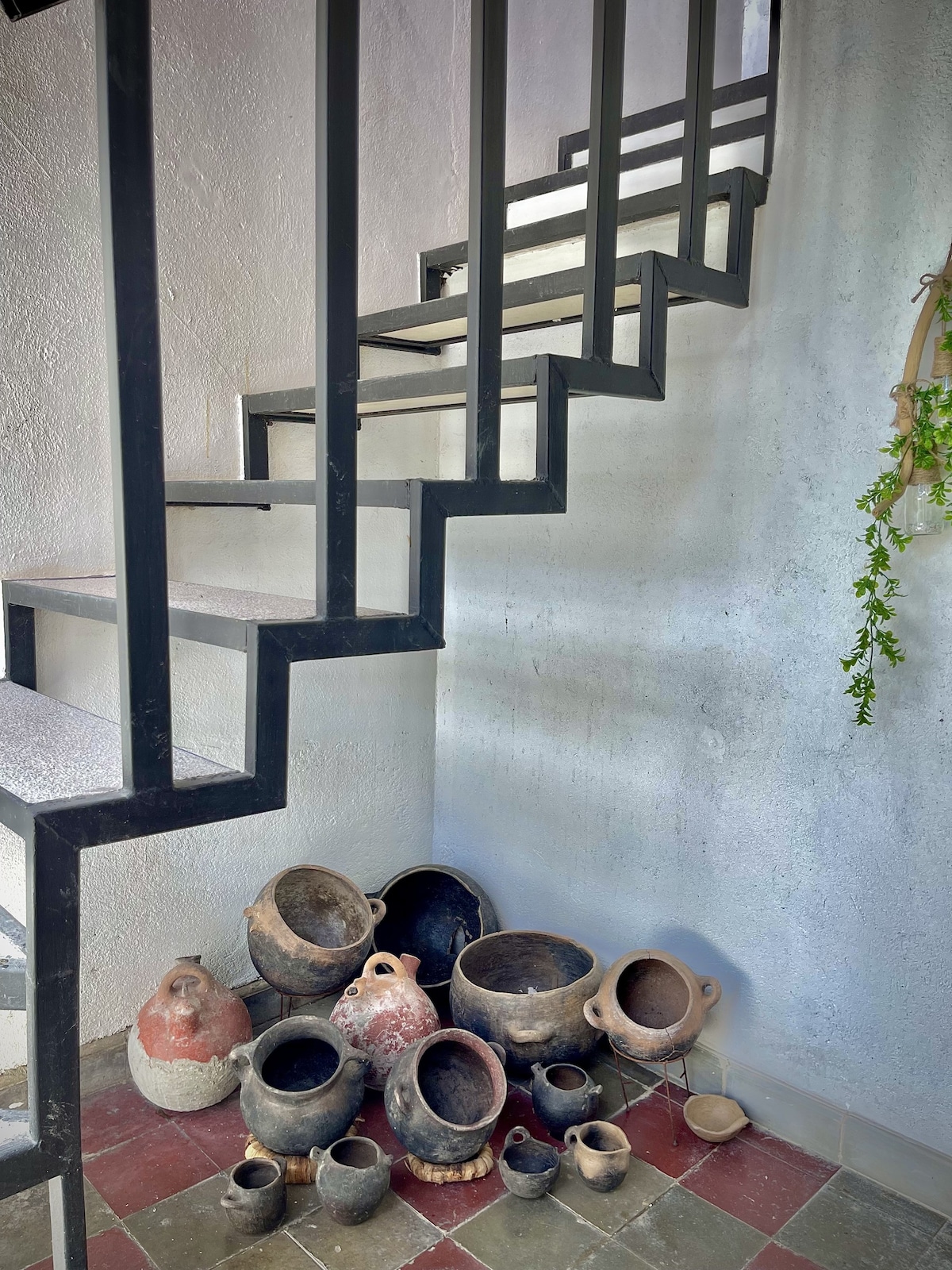 A modern staircase with a sleek black railing ascends alongside a textured white wall. At the base, a collection of traditional pottery in various sizes is displayed on the floor, showcasing earthy tones and unique shapes.