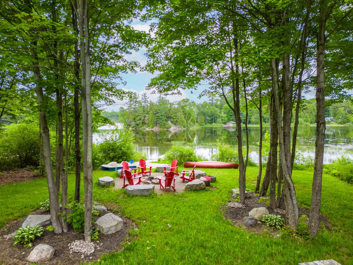 A serene outdoor gathering space is nestled among trees, featuring a circular arrangement of vibrant red chairs around a stone fire pit. The lush green grass extends towards calm waters reflecting the surrounding nature, complemented by a few colorful kayaks resting nearby.