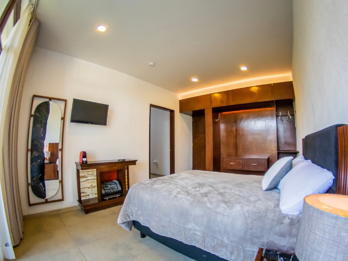 A spacious bedroom features neutral-colored walls and flooring. A large window allows natural light, complemented by warm overhead lighting. A comfortable bed is situated against a wooden headboard, with flat-screen TV mounted on the wall. Storage and hanging space are provided in wooden cabinetry.