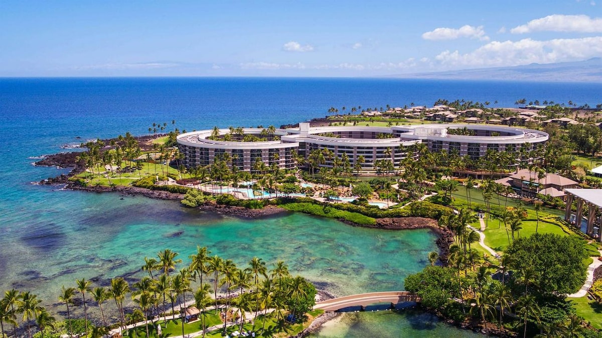 Aerial view of the resort showcases an expansive coastline bordered by vibrant blue waters. Lush green landscaped grounds are accented by palm trees, with multiple pools visible on the property. The architecture of the building is modern and curved, complementing the natural surroundings.