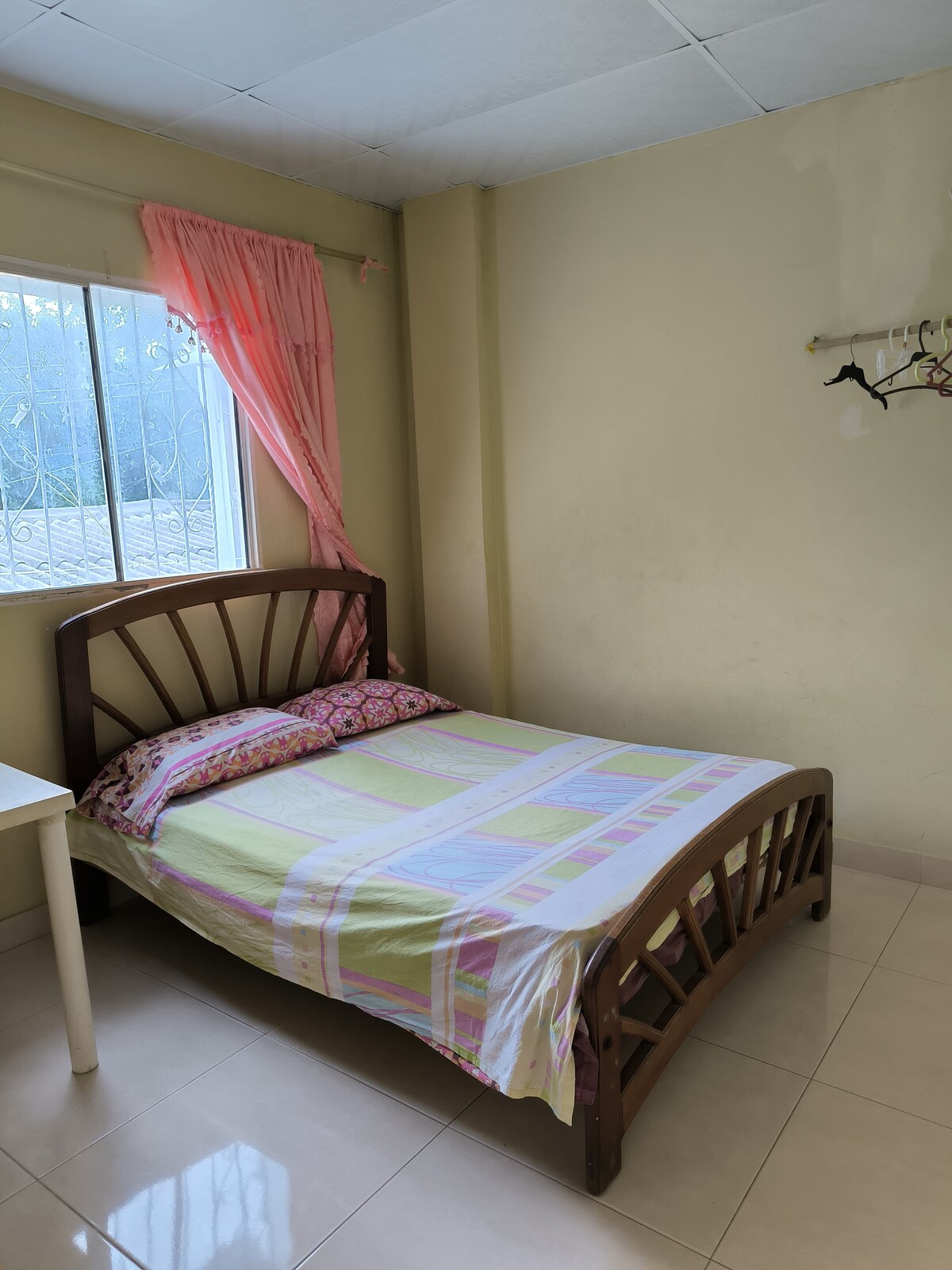 A simple sleeping area features a wooden bed with a patterned blanket and a decorative pillow. A window with a light pink curtain allows natural light to fill the room, while a small table is positioned beside the bed.
