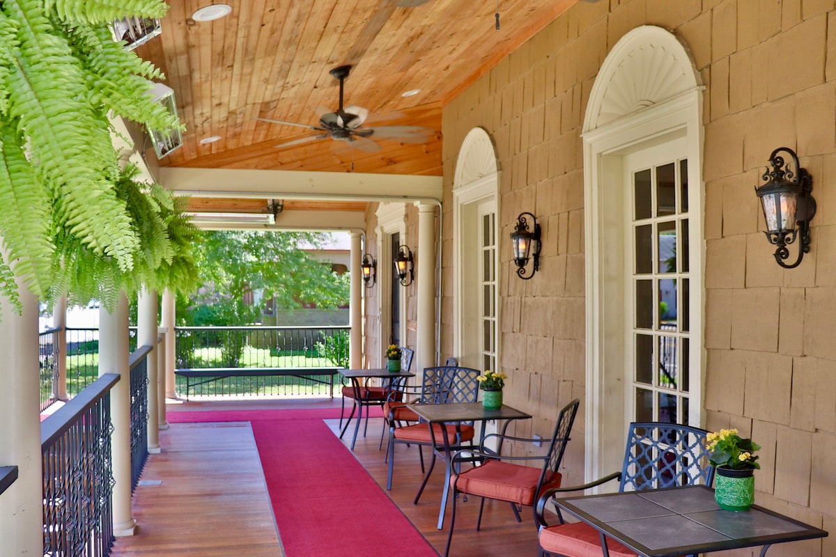 An expansive porch features comfortable seating arranged around small tables. Lush ferns hang from the ceiling, and decorative lanterns are mounted on the walls. A welcoming red carpet runs along the floor, creating a cozy environment for relaxation.
