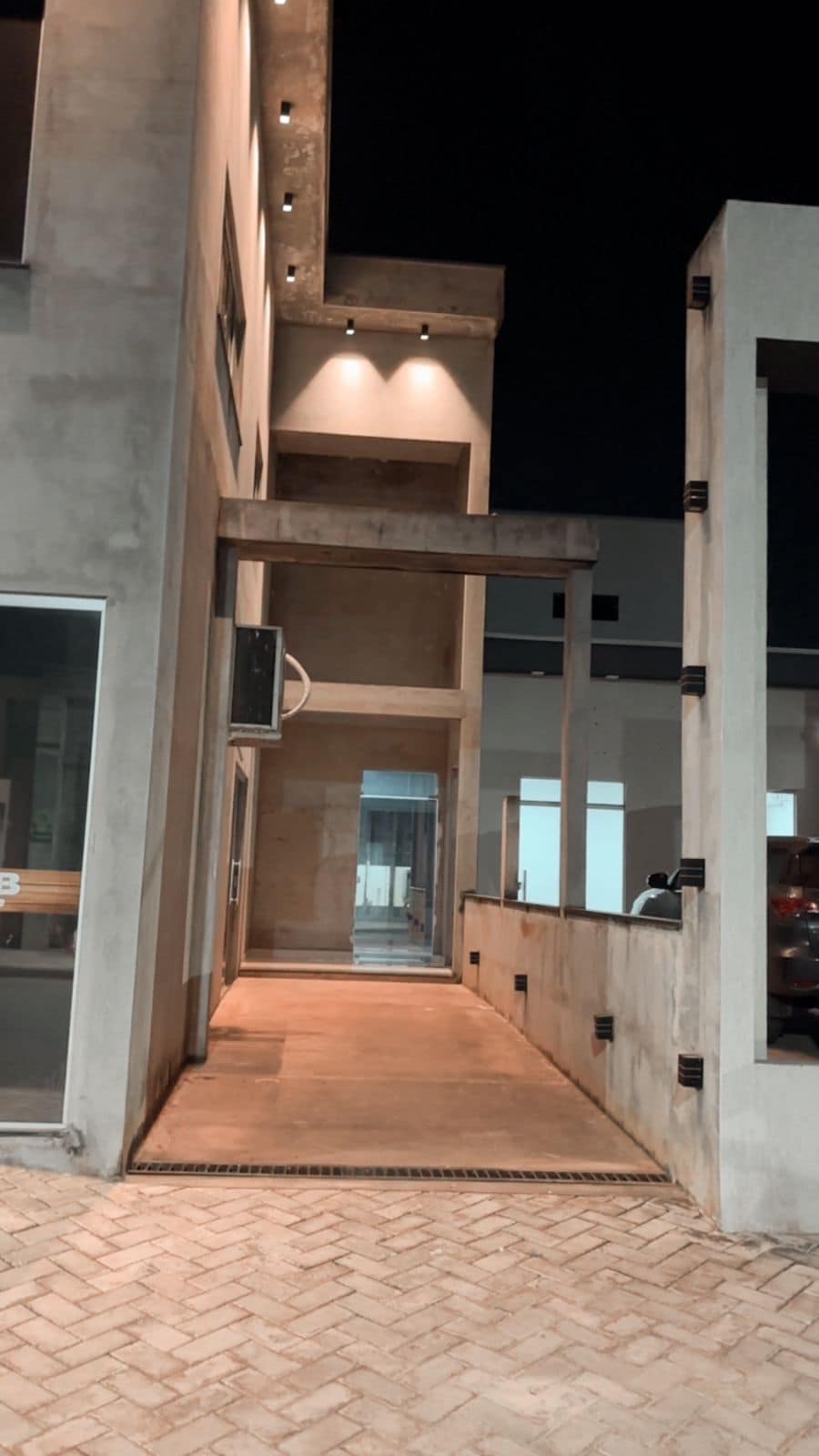 A wide pathway leads to the entrance of a modern building, framed by concrete walls. Soft lighting highlights the contemporary architecture, while large windows allow glimpses of the interior spaces. The pathway is surrounded by minimalist landscaping, enhancing the structured design.