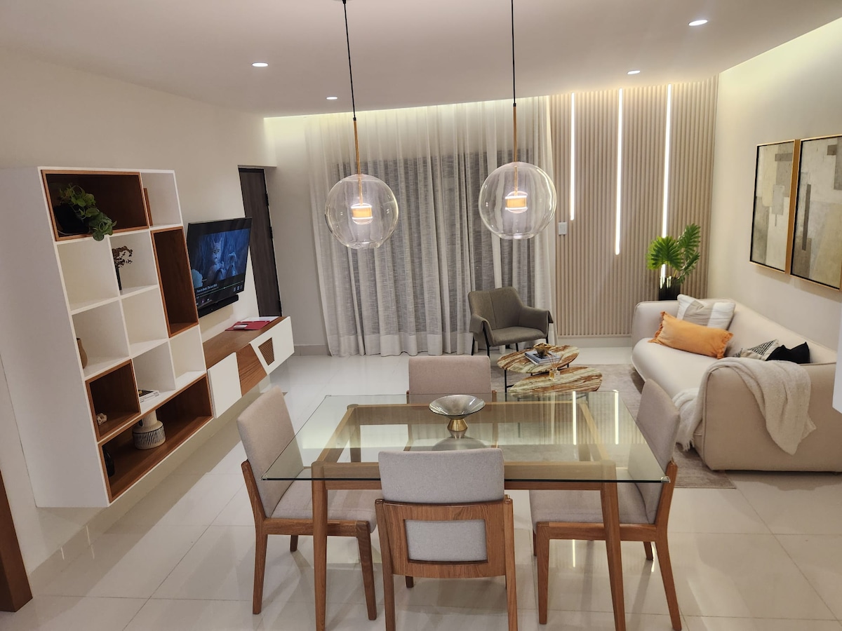 A spacious living area is depicted, featuring a glass dining table surrounded by four wooden chairs. A light-colored sofa is positioned nearby, complemented by decorative pillows. Soft lighting from pendant fixtures creates a calming ambiance, while large windows are dressed with sheer curtains.