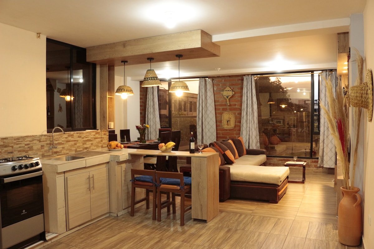 An open living space is presented, featuring a modern kitchen with stainless steel appliances and a dining area with wooden furniture. A comfortable sofa is positioned adjacent to large windows, allowing natural light to illuminate the space. Decorative elements add warmth to the atmosphere.