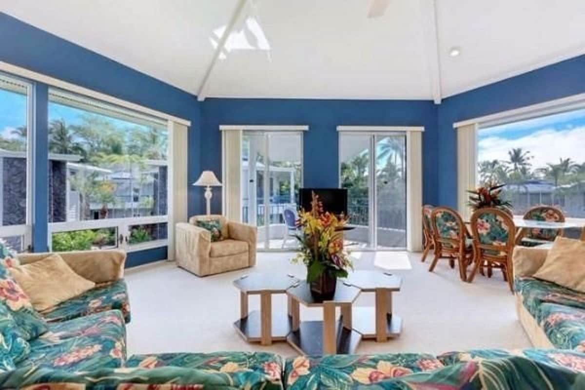A spacious living area features large windows that allow natural light to fill the room. Tropical-patterned sofas are arranged around a coffee table, while a dining set sits nearby. A ceiling fan is present, and lush greenery can be seen outside.