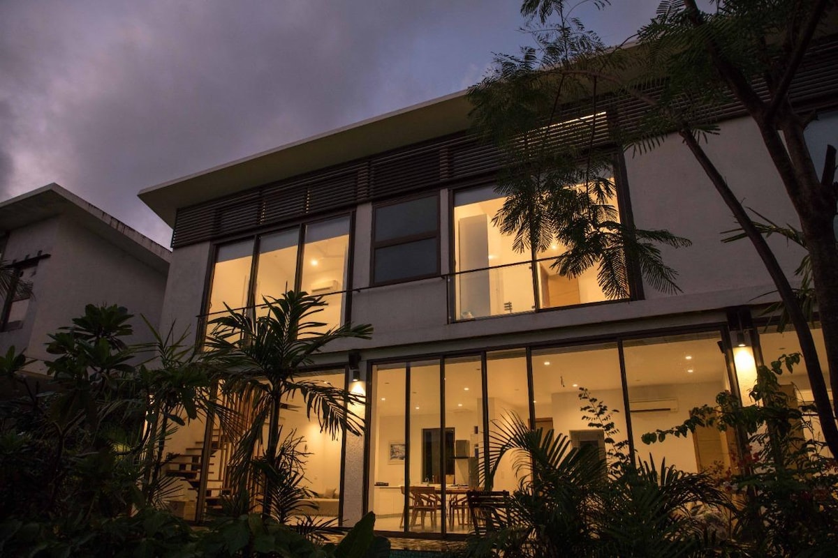 The villa's exterior is showcased at dusk, with large glass doors revealing warm interior lighting. Lush greenery frames the space, enhancing the sense of tranquility. The modern architecture emphasizes clean lines and natural light, inviting guests to explore the relaxing outdoor environment.
