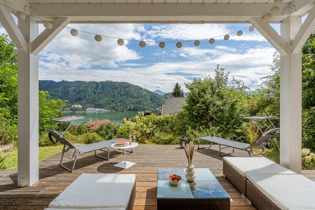 A spacious terrace features comfortable seating options, including loungers and a wicker couch. The area is framed by greenery and provides a panoramic view of the mountains and the nearby Ossiacher See, creating an inviting outdoor space for relaxation.