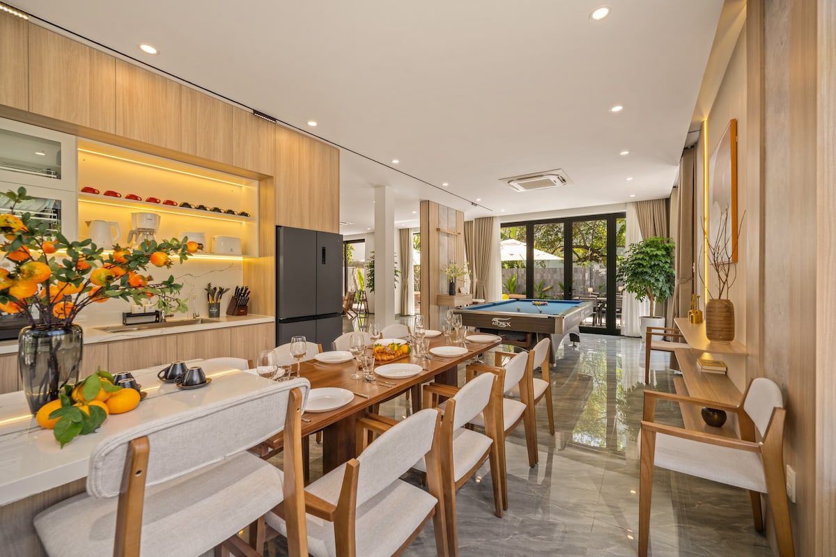 The dining area is highlighted by a long wooden table set for multiple guests, surrounded by six cushioned chairs. A modern kitchen with sleek cabinetry is visible, along with a pool table and large glass doors leading to an outdoor patio filled with greenery.