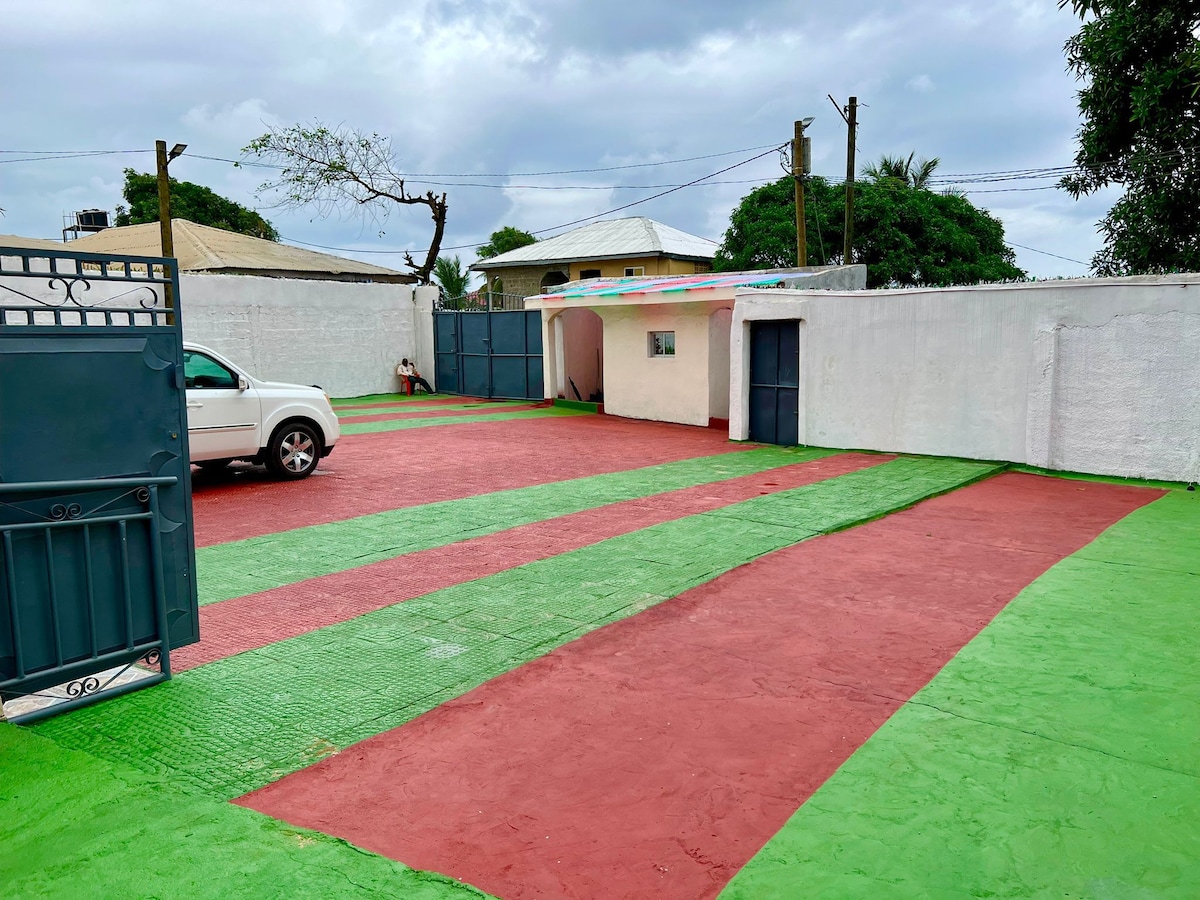 A spacious outdoor parking area is designed with alternating red and green sections. A white vehicle is parked near a light-colored building with a door visible. Surrounding the area are green grass patches and nearby trees, creating a pleasant outdoor environment.