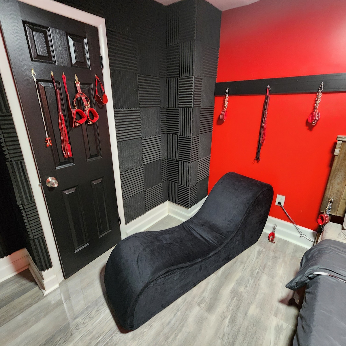 A playroom corner is highlighted by a sleek, dark lounge piece against a striking red and black wall. Various accessories hang on the wall, adding character, while textured panels provide soundproofing. A door with a dark finish is visible, maintaining a cohesive design.