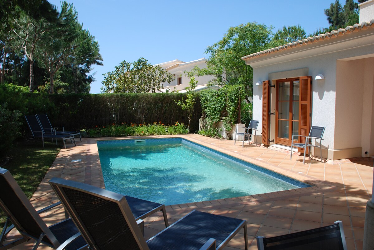 A private pool is set within a terraced area surrounded by well-maintained gardens. Several lounge chairs are positioned on the patio, providing a space for relaxation. Bright sunlight illuminates the clear water, creating a refreshing ambiance.