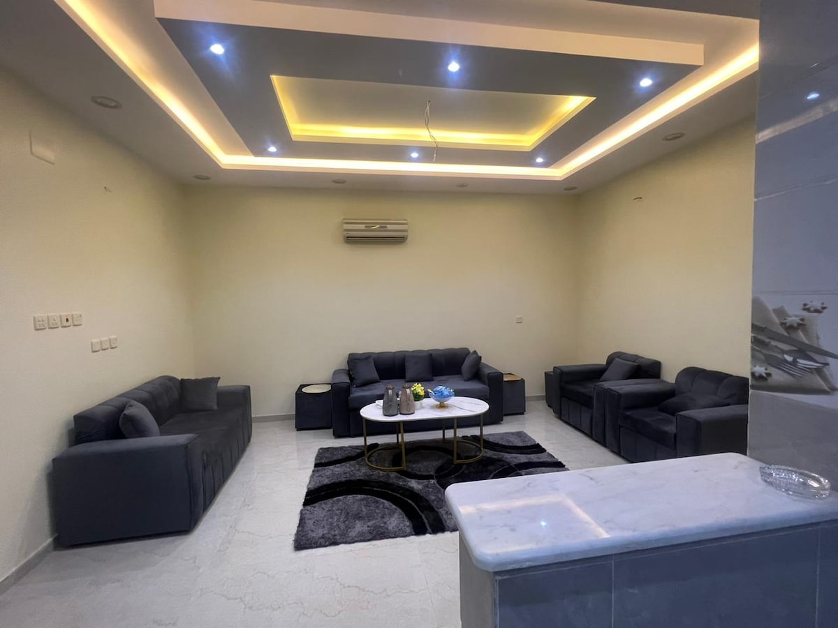 A spacious living area features comfortable seating arrangements with dark sofas and armchairs, placed around a circular coffee table. A patterned rug covers the floor, and recessed lighting illuminates the ceiling, creating a modern and inviting environment.