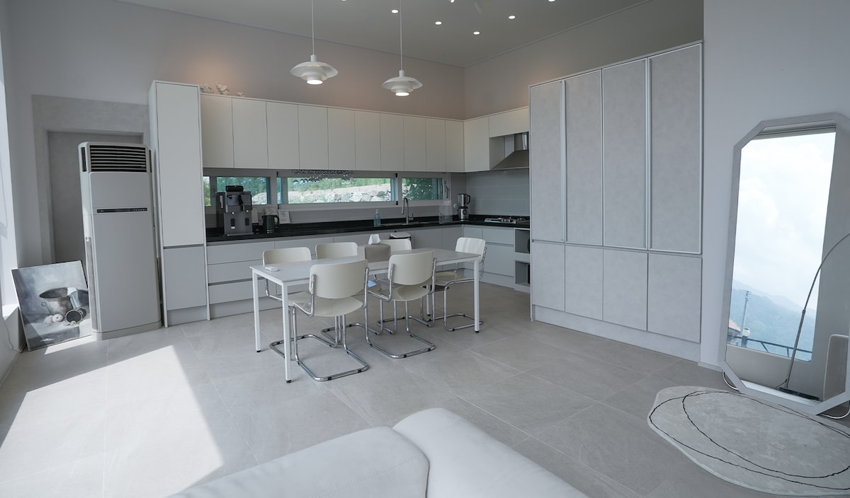A modern kitchen is showcased with sleek cabinetry and a spacious dining area. A large window provides a view of the outdoors, while a dining table is surrounded by four chairs. Additional appliances, including a refrigerator and a stove, are neatly arranged within the space.