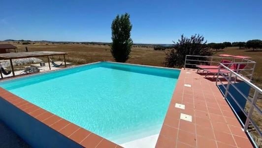 A rectangular pool is surrounded by a tiled patio, featuring two lounge chairs. Expansive views of the natural landscape can be seen in the background under a clear blue sky.