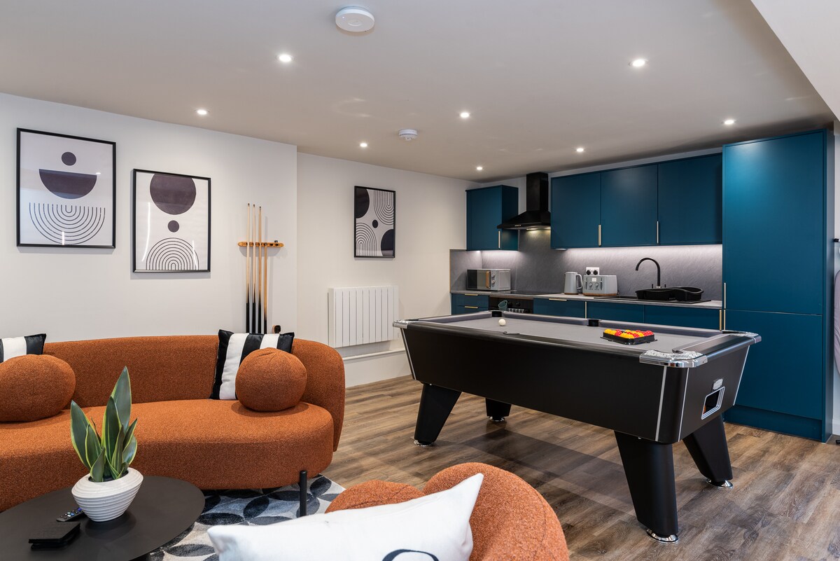The living area features a modern design with a vibrant orange couch and a black pool table at the center. Stylish framed artwork adorns the walls, and a well-equipped kitchen with blue cabinetry is visible in the background, offering a blend of comfort and entertainment.