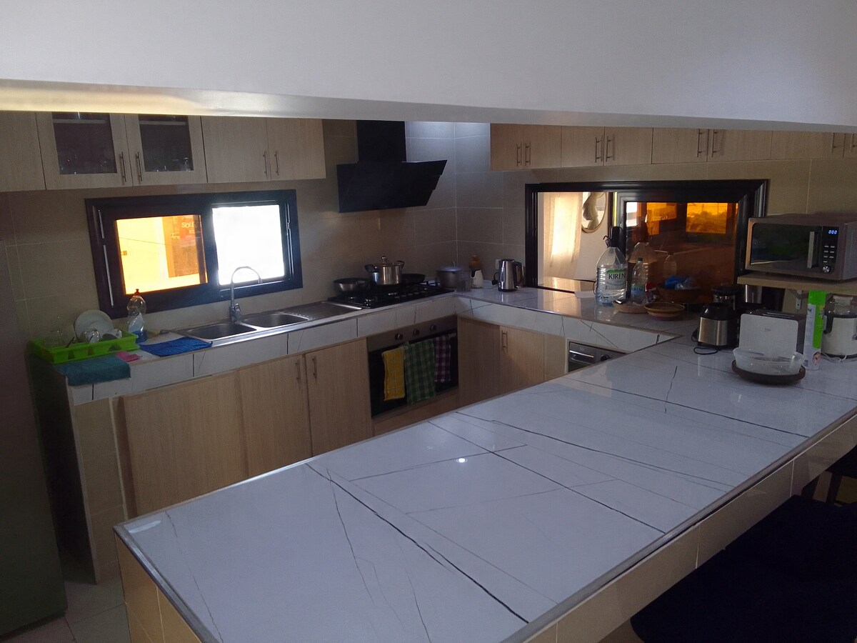 A modern kitchen features a central island with a marble countertop and ample workspace. Equipped with cabinetry and essential appliances, including a microwave and stove. Natural light comes through two windows, illuminating the clean and functional layout.