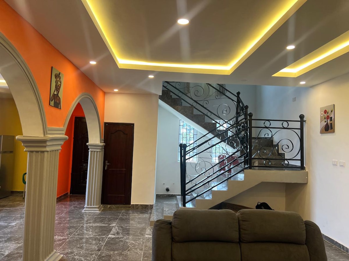 A spacious interior is accented by an orange feature wall, creating a vibrant atmosphere. Curved archways lead to a staircase with decorative black railings. The floor features glossy tiles, reflecting light from recessed ceiling lights above.