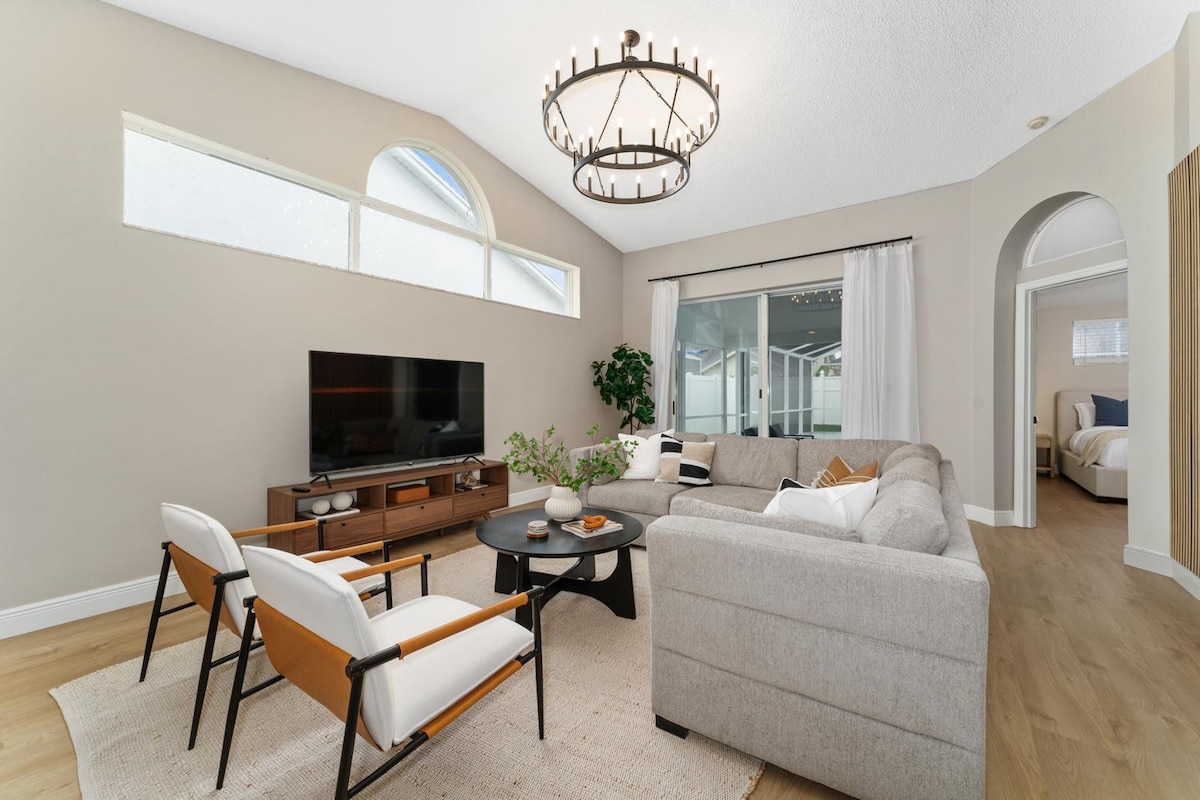 A modern living area is presented, featuring a light-colored sofa and two stylish chairs arranged around a circular table. A flat-screen television is mounted on the wall opposite a large window that allows natural light to fill the space. Neutral tones create a calming ambiance.