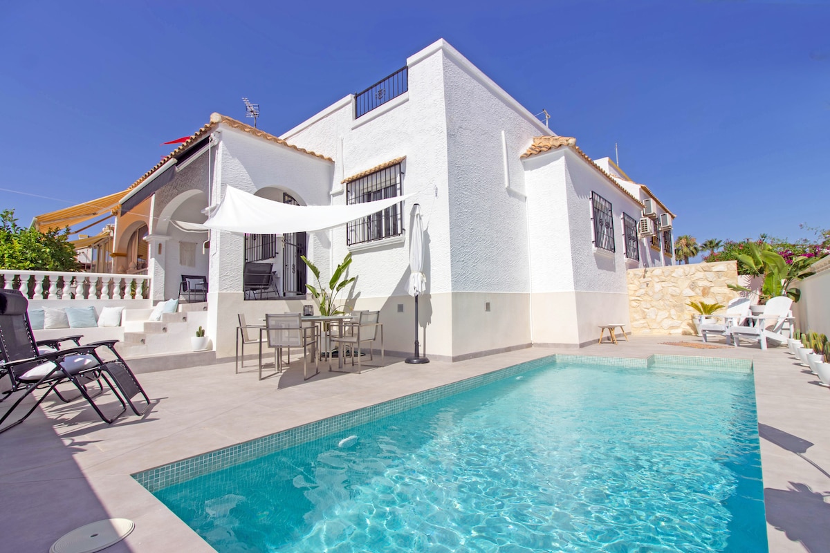 A bright holiday home is showcased with a private swimming pool surrounded by a spacious terrace. Sun loungers are positioned around the pool, and a shaded dining area is visible. The white exterior of the house contrasts with the blue water, creating a welcoming environment.