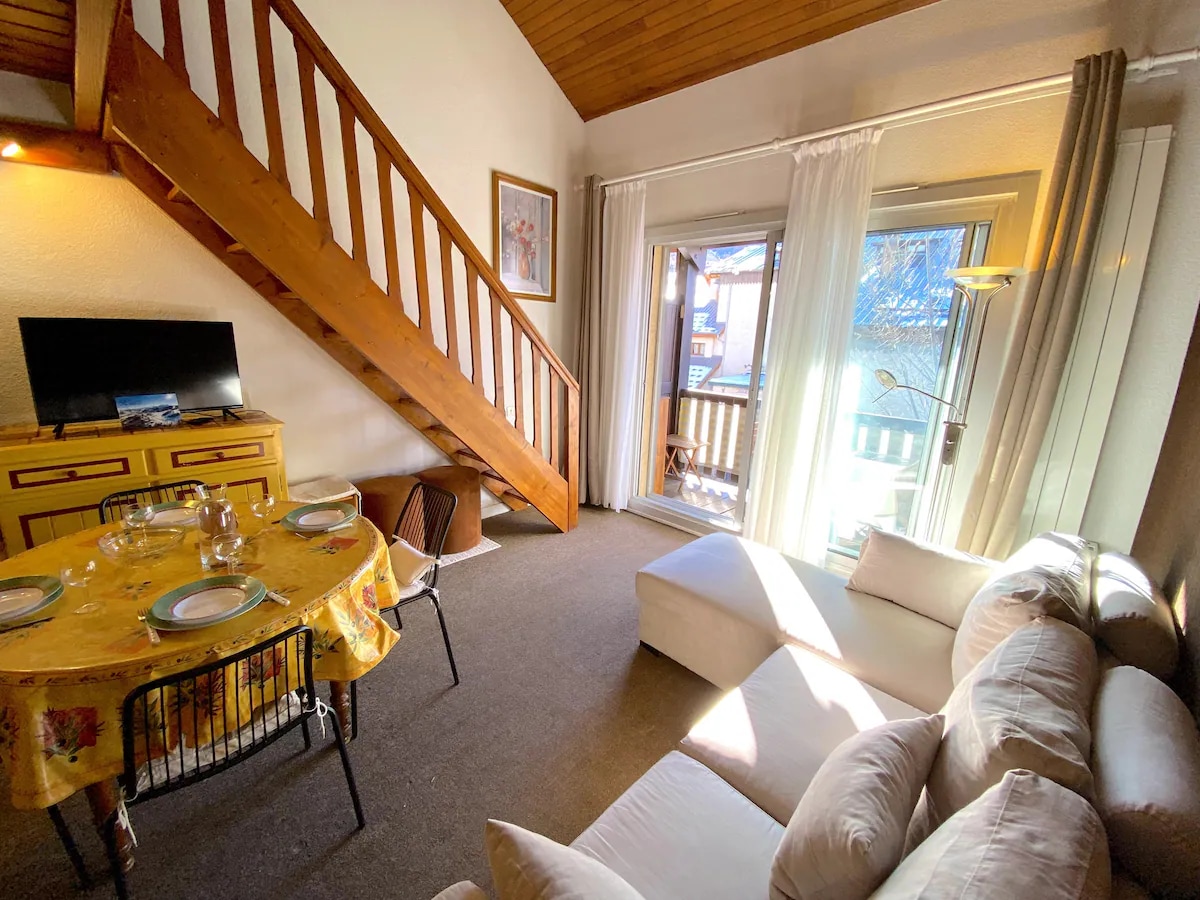 A welcoming living area features a light-colored sofa and a round dining table set for four. Natural light streams through tall windows, illuminating the space. A wooden staircase leads to an upper level. A television is positioned on a colorful cabinet.