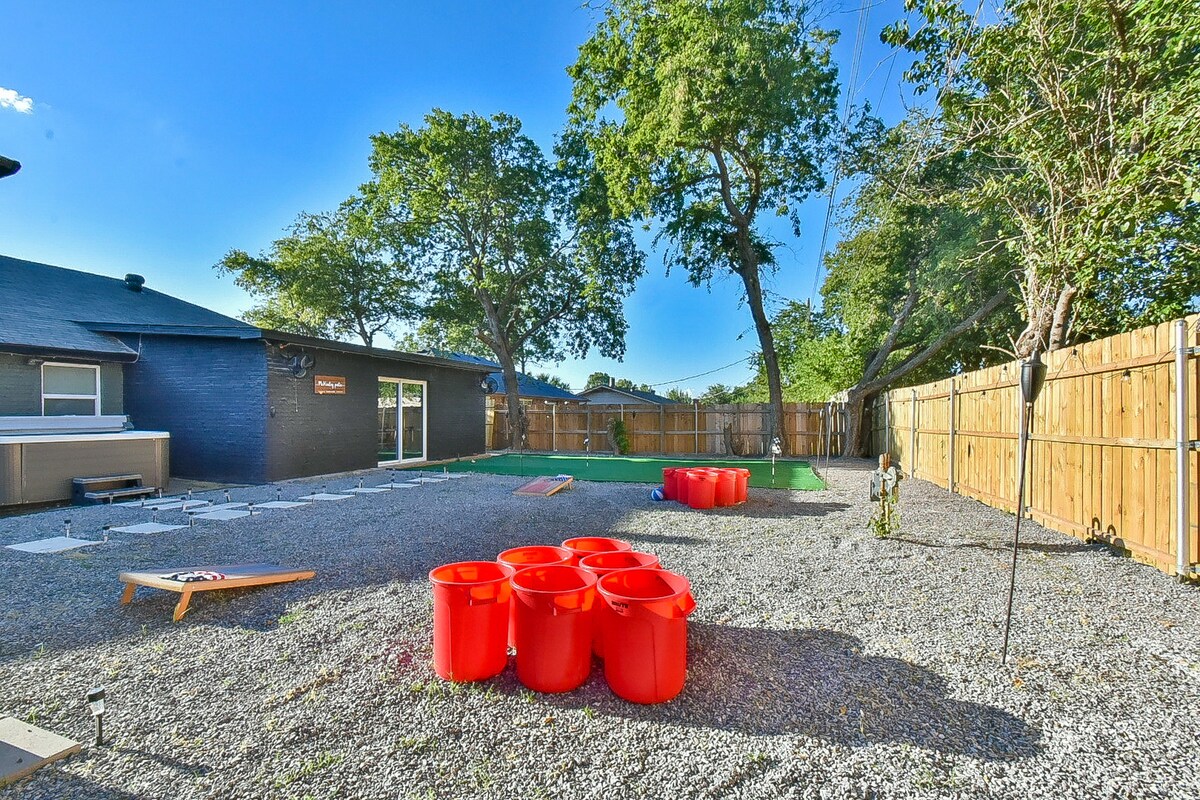 An outdoor area features a spacious gravel surface, bordered by a wooden fence. Bright orange yard pong cups are arranged on the ground, and a mini golf course is visible in the background. Lush trees provide shade in this recreational space.