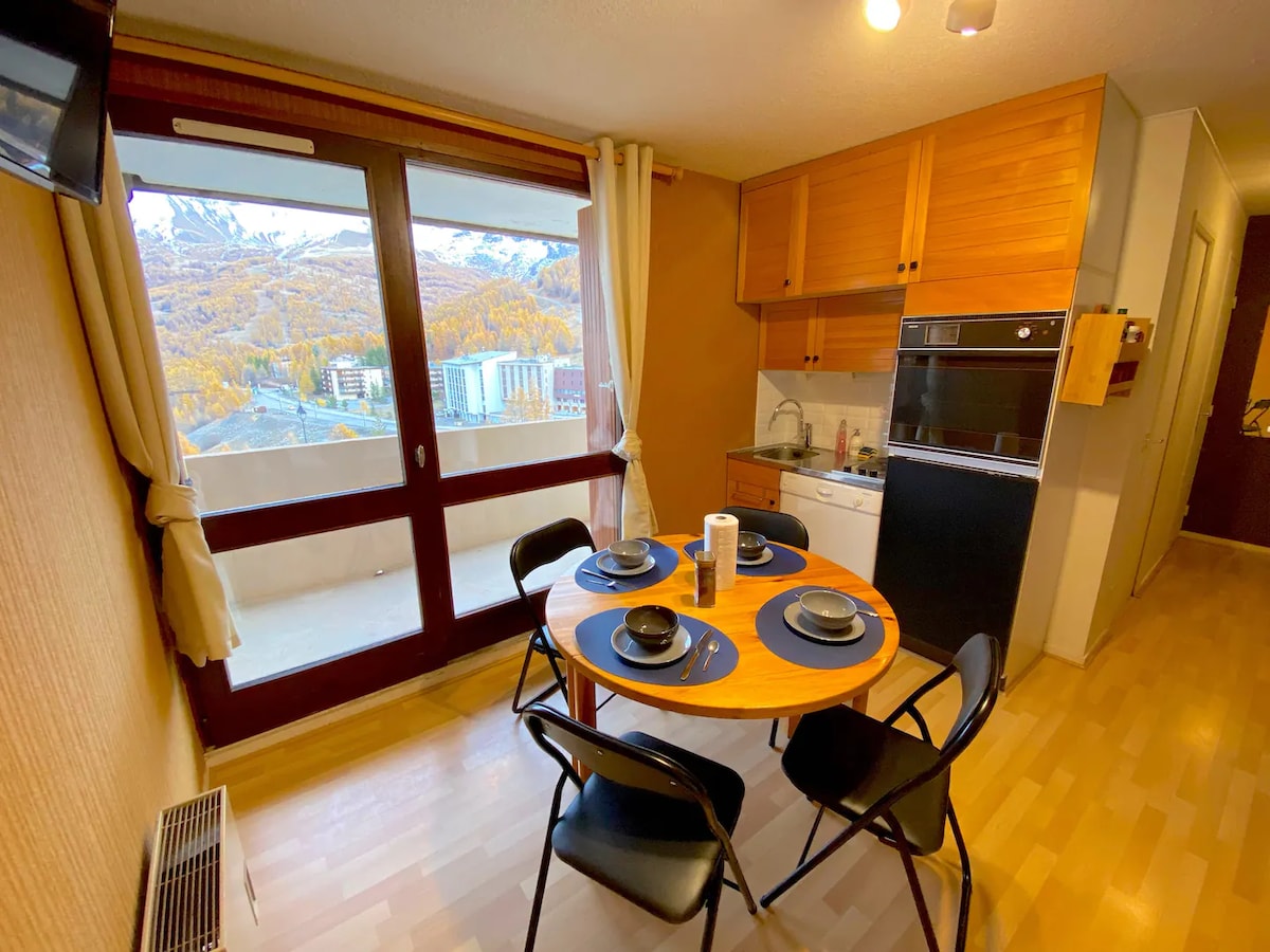A dining area features a round table set for four, accompanied by black chairs. Large windows showcase a beautiful view of the surrounding mountains, while the kitchen is equipped with essential appliances, including a dishwasher. Natural light enhances the warm wooden tones of the cabinetry.
