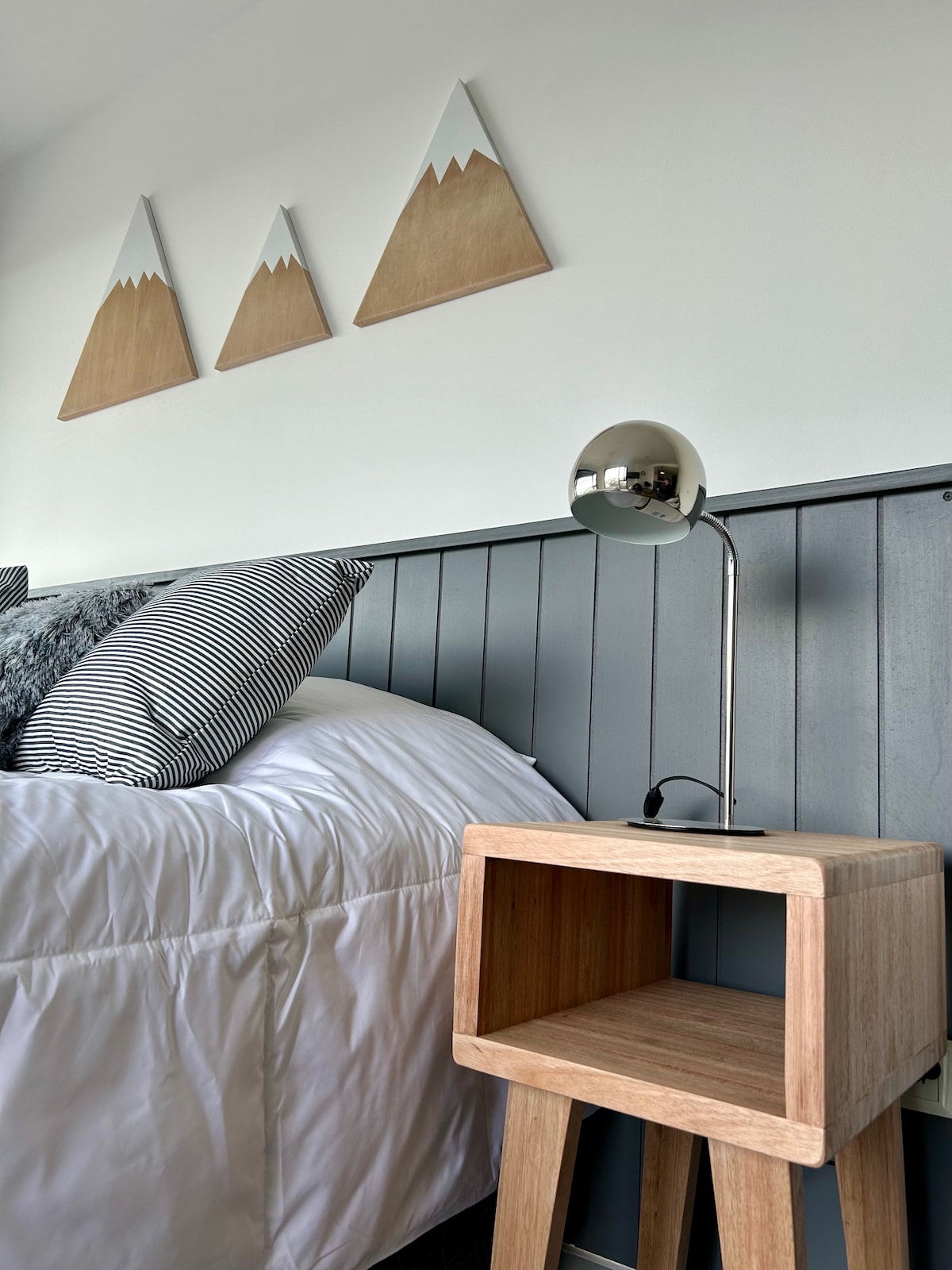 A well-appointed sleeping area features a spacious bed with a white comforter and patterned pillows. A light wood nightstand holds a modern silver lamp. Three triangular mountain decorations are displayed on the wall above, complementing the neutral color scheme.