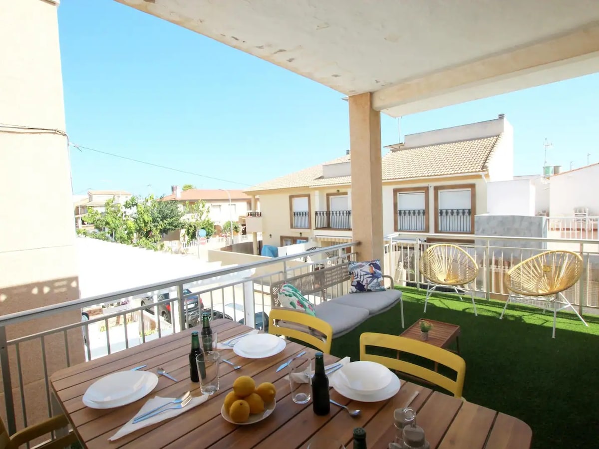 An outdoor terrace features a dining table set with plates and a bowl of lemons, surrounded by wooden chairs. Two lounge chairs are positioned nearby. The area is bright and airy, with views of neighboring buildings and clear blue skies.