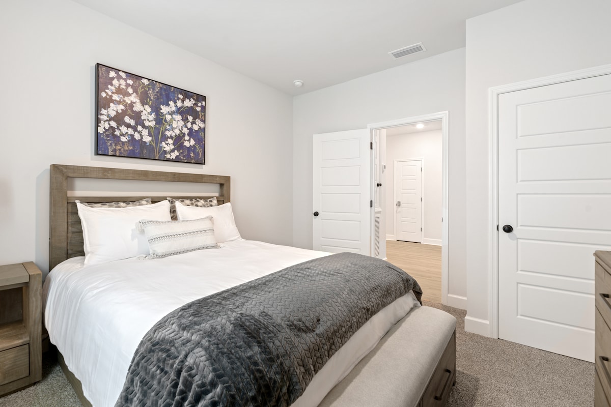 Dormitorio sereno con una acogedora cama tamaño queen con sábanas suaves y un televisor, flanqueado por modernas mesas auxiliares. Los tonos neutros de la habitación y las obras de arte de buen gusto crean un ambiente tranquilo, perfecto para relajarse