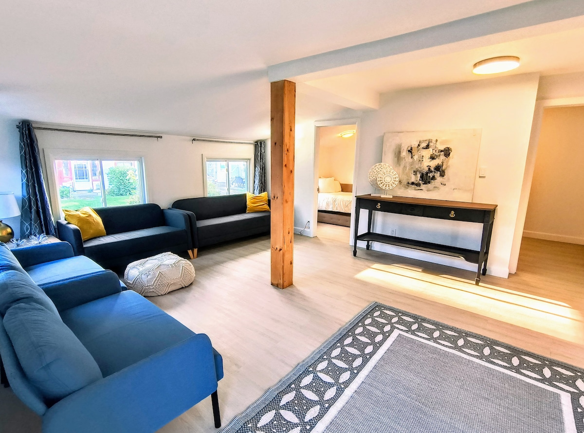 A bright living room is visible, featuring two plush sofas in varying shades of blue, a decorative pouf, and a sleek black console table. Large windows allow natural light to fill the space, highlighting the modern décor and clean lines. A hint of a cozy bedroom is seen in the background.