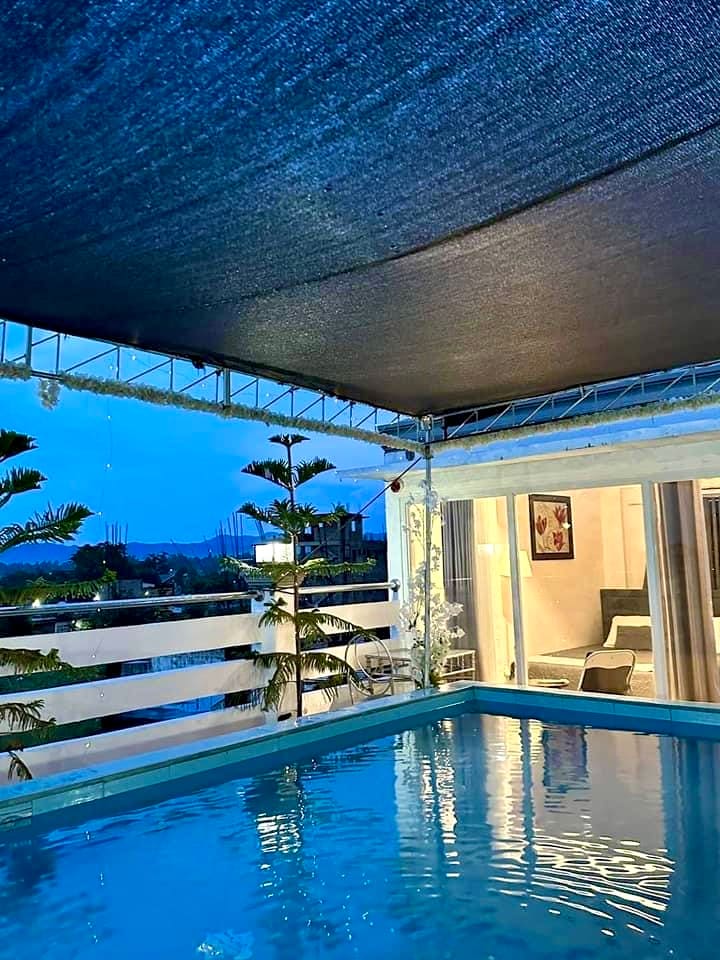 A serene plunge pool is positioned on a roof deck, surrounded by lush greenery and soft lighting. The evening sky is visible through the open structure, with a cozy interior space seen in the background, featuring neutral tones and contemporary furnishings.