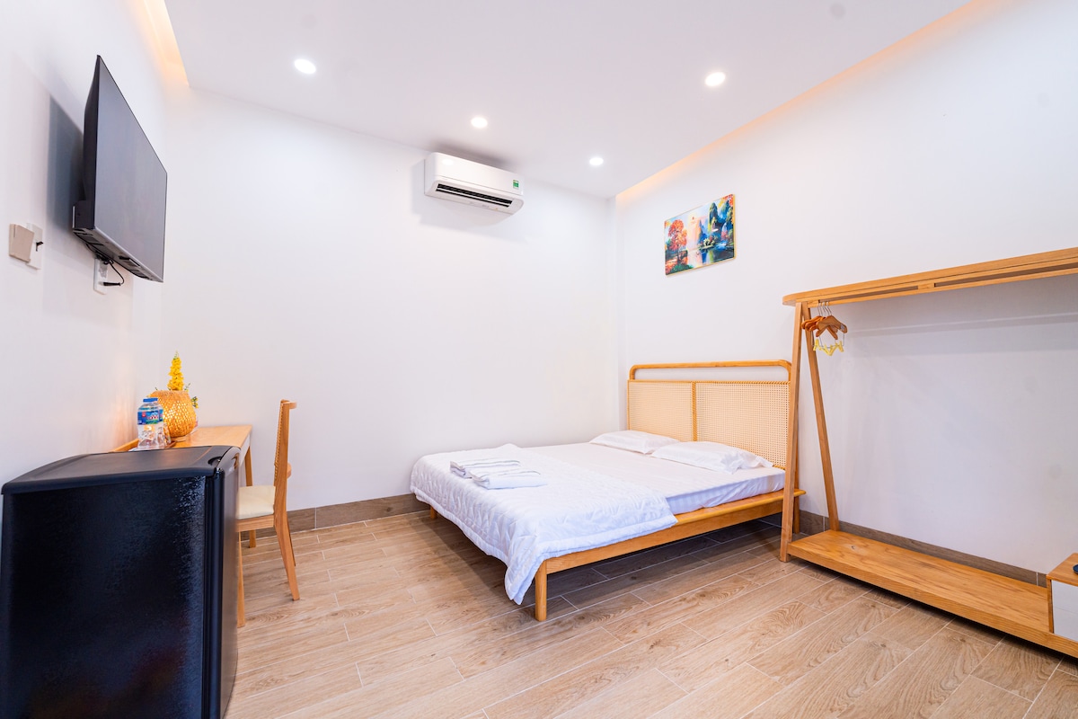 A serene sleeping area features a spacious bed with neatly arranged linens. A wall-mounted television is present opposite the bed. An air conditioning unit and a piece of wall art add subtle touches. A wooden clothing rack and a small desk enhance the functional space.