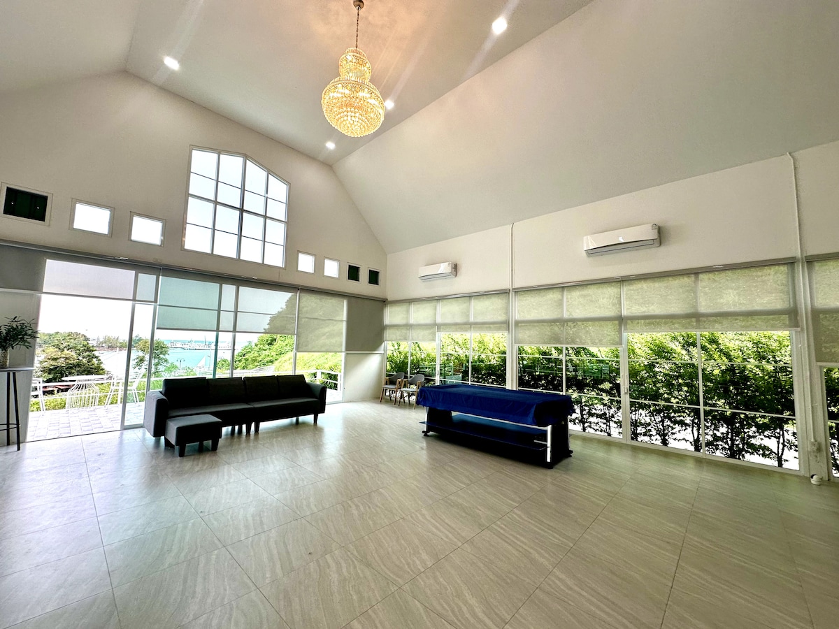 A spacious common area features high ceilings and large windows, allowing natural light to fill the room. Modern seating arrangements include a dark sofa and a blue bench, while air conditioning units provide comfort. Lush greenery is visible through the windows, enhancing the serene environment.