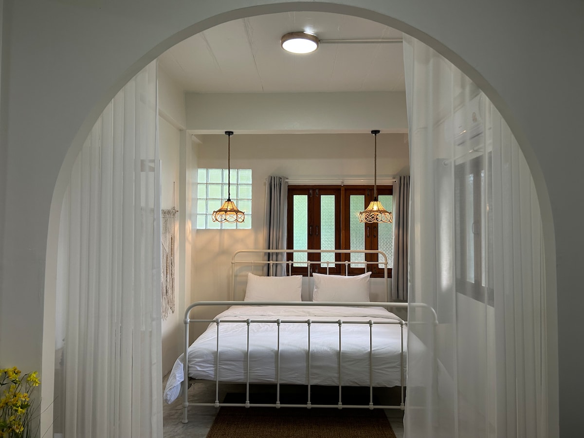 A bright sleeping area features a spacious bed framed by soft curtains. Two hanging light fixtures provide gentle illumination. Large windows on the opposite side allow natural light to enter, while neutral tones create a calming ambiance throughout the space.