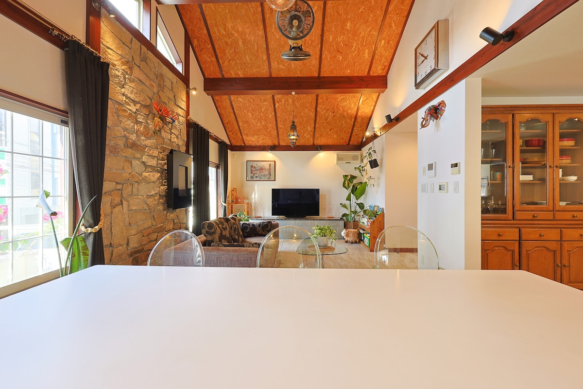 A spacious living area is highlighted by a high ceiling with wooden beams and stone accents. Natural light streams through large windows, illuminating the cozy seating area with plants, while a dining space features a modern, white table in the foreground.