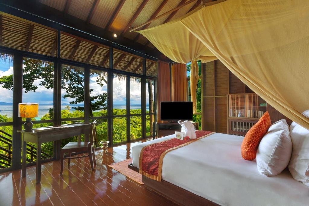 A spacious bedroom features a large bed draped with a sheer canopy. Floor-to-ceiling windows offer views of the lush, green landscape. A small desk is positioned near the window, and natural light fills the room, highlighting the warm wood tones and decorative accents.