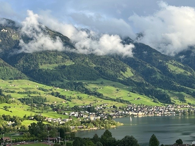 A panoramic view captures a tranquil landscape of lush green hills and scattered homes along the water's edge. Majestic mountains rise in the background, partially shrouded in clouds, reflecting a serene and natural environment.