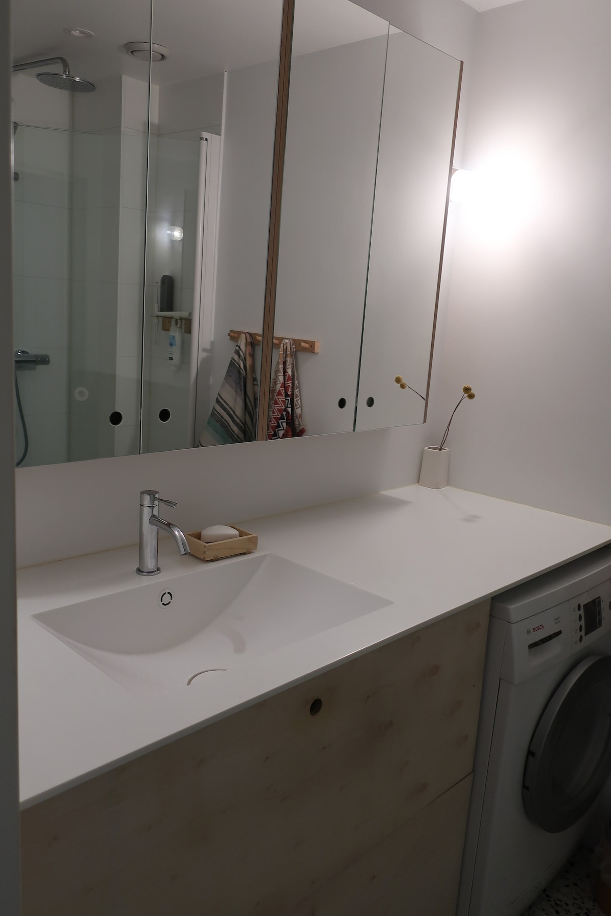 The bathroom features a modern sink area with a spacious countertop, accompanied by a washing machine. A large mirror above the sink reflects the clean lines of the design. Simple decor elements, including a soap dish and a flower, add subtle touches.